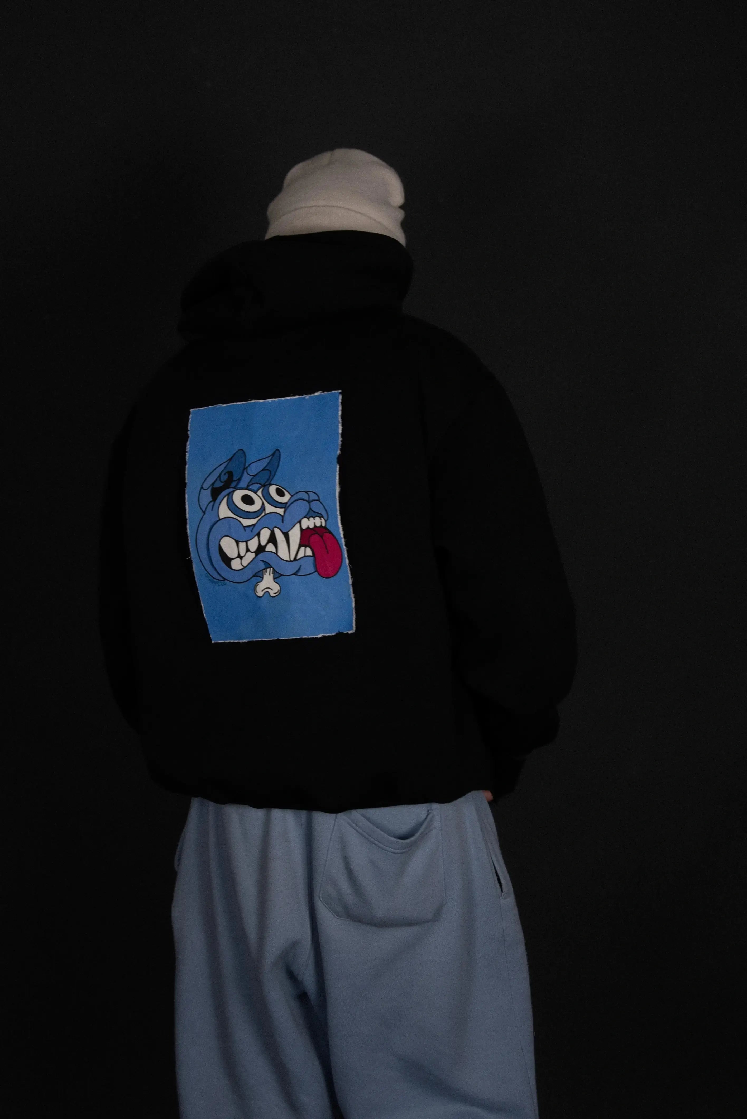 Felpa nera con patch magnetica Cryptic Mad Dog blu sul retro, indossata da modello con cappuccio e pantaloni chiari, stile streetwear personalizzabile.
